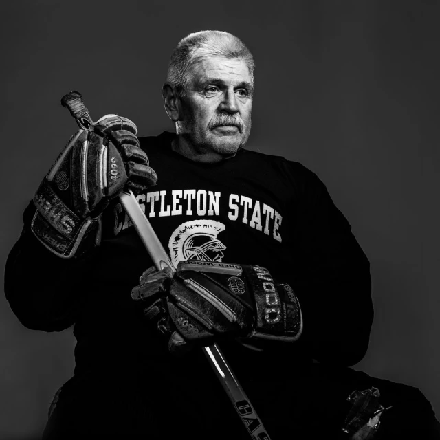 Monochrome portrait of older man in 'Castleton State' sweatshirt, holding hockey stick with gloves; stark lighting, studio setting.