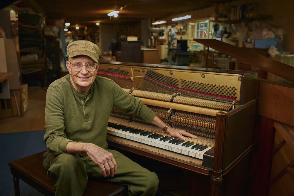Elderly man smiles, sits at upright piano in workshop, wearing green clothing and a cap, showcasing musical instrument repair.