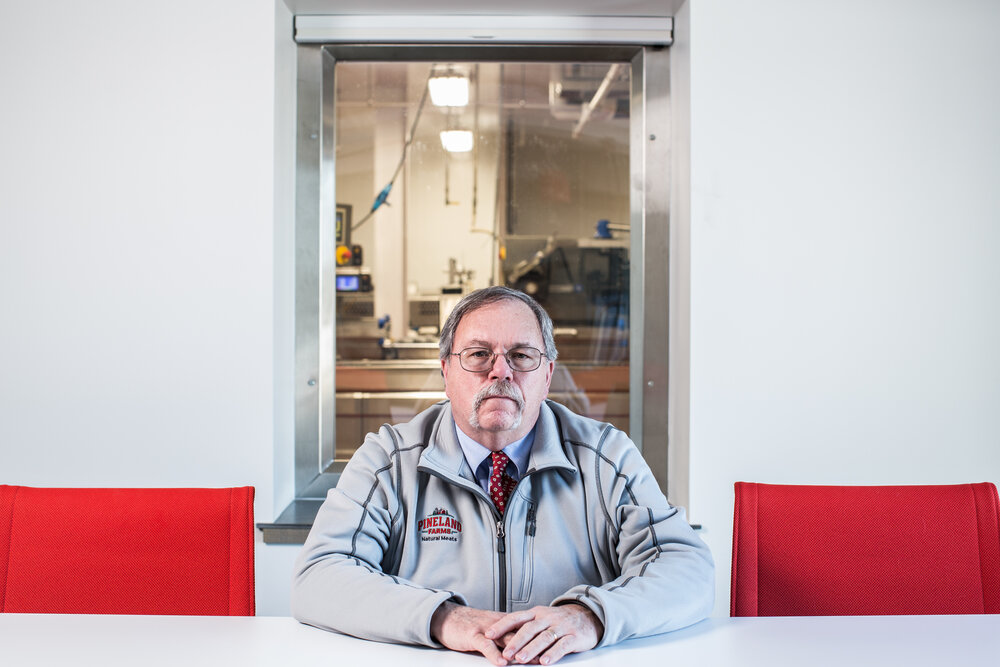 Portrait of a mature man in a grey jacket sitting at a white table in front of a window with an industrial background.