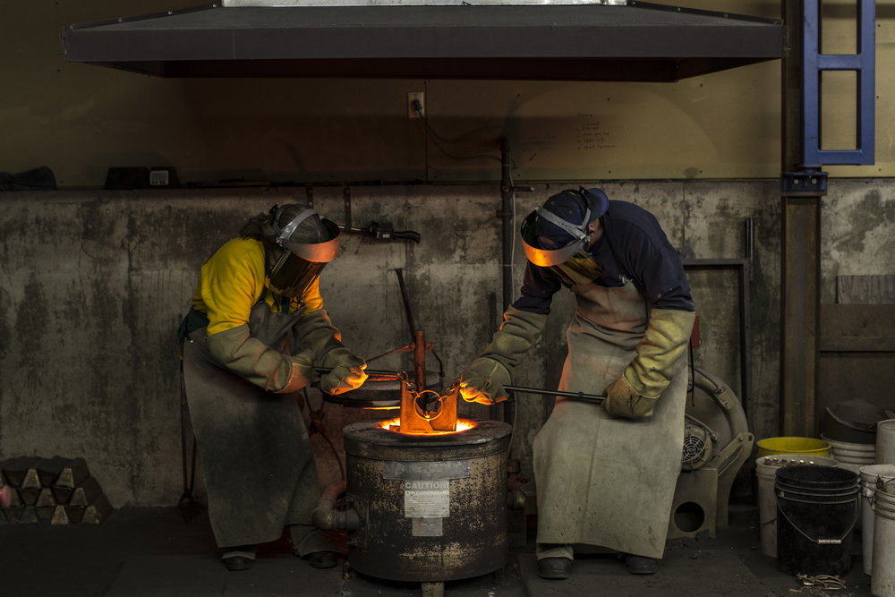 Two industrial workers in protective gear manipulate molten metal at a foundry, with sparks and intense heat visible in a dimly lit factory.