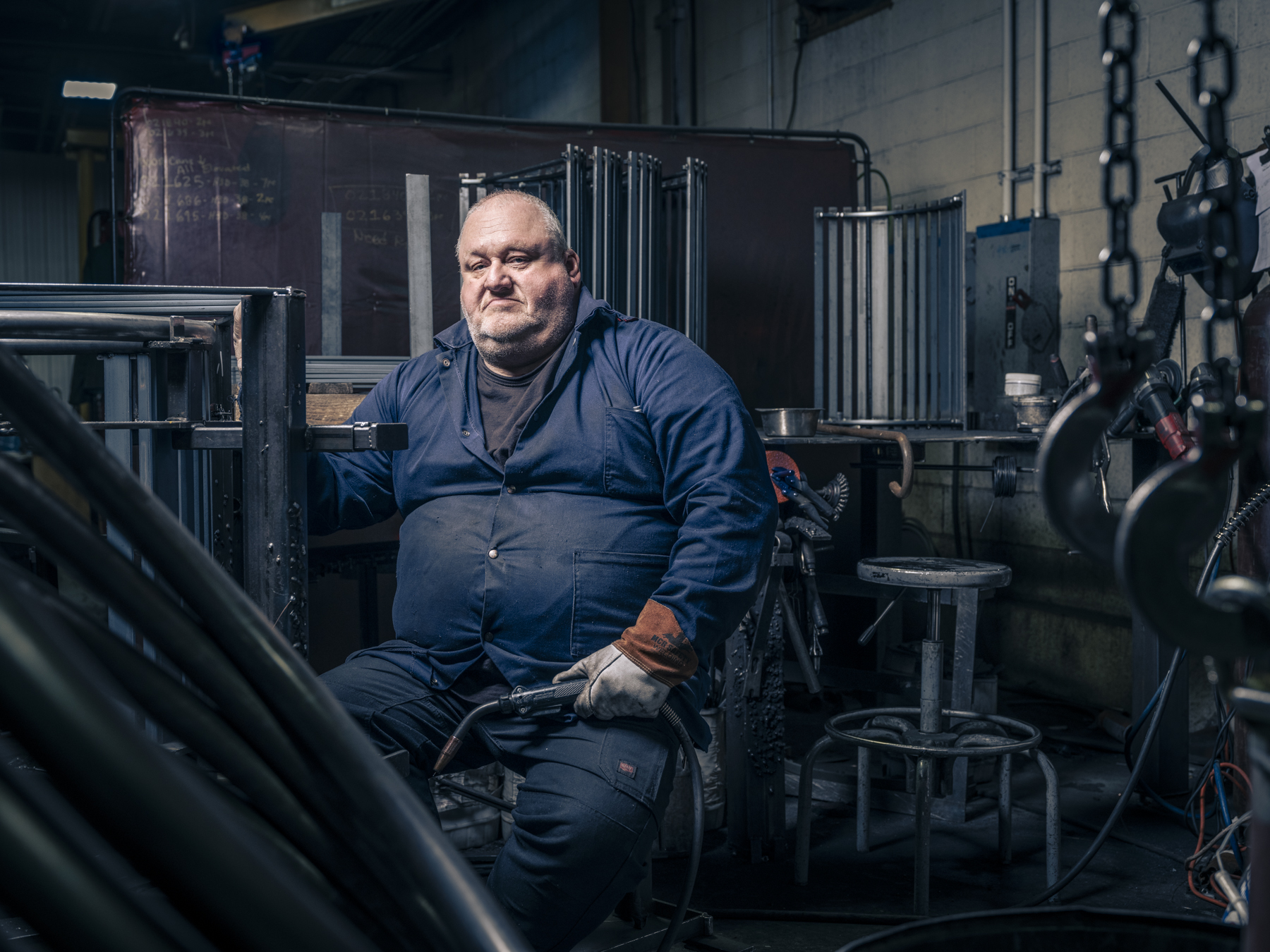 Man in navy work suit posing indoors with welding equipment. He sits on a stool surrounded by metal tubes in a dimly lit factory.