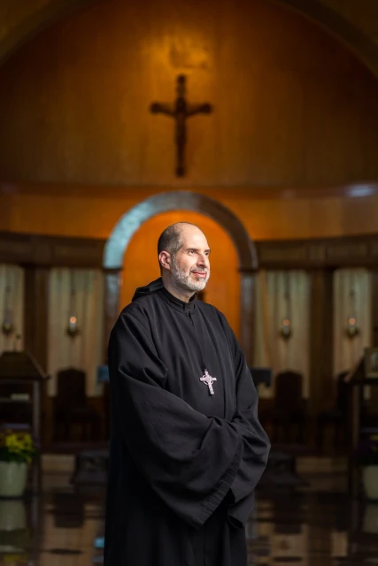 Portrait of a middle-aged man wearing a black robe with a cross necklace standing in a church with golden walls and a crucifix.