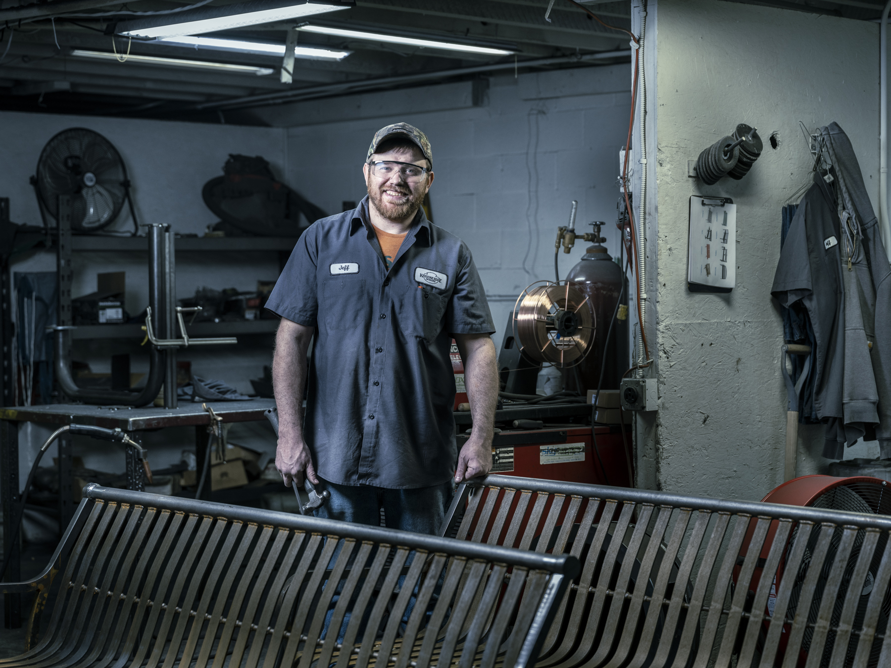 Man in work shirt and safety glasses poses with metal bench in an industrial setting with welding equipment and tools in the background.