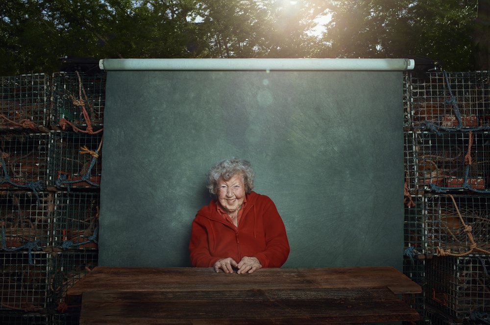 Elderly woman with white hair and a red sweater seated at a wooden table in front of a green screen backdrop and stacked crab traps.