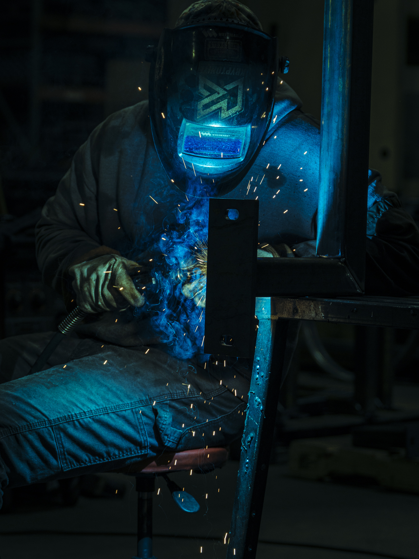 Person wearing a welding mask works with blue sparks flying in a dark industrial setting; metal frame visible.