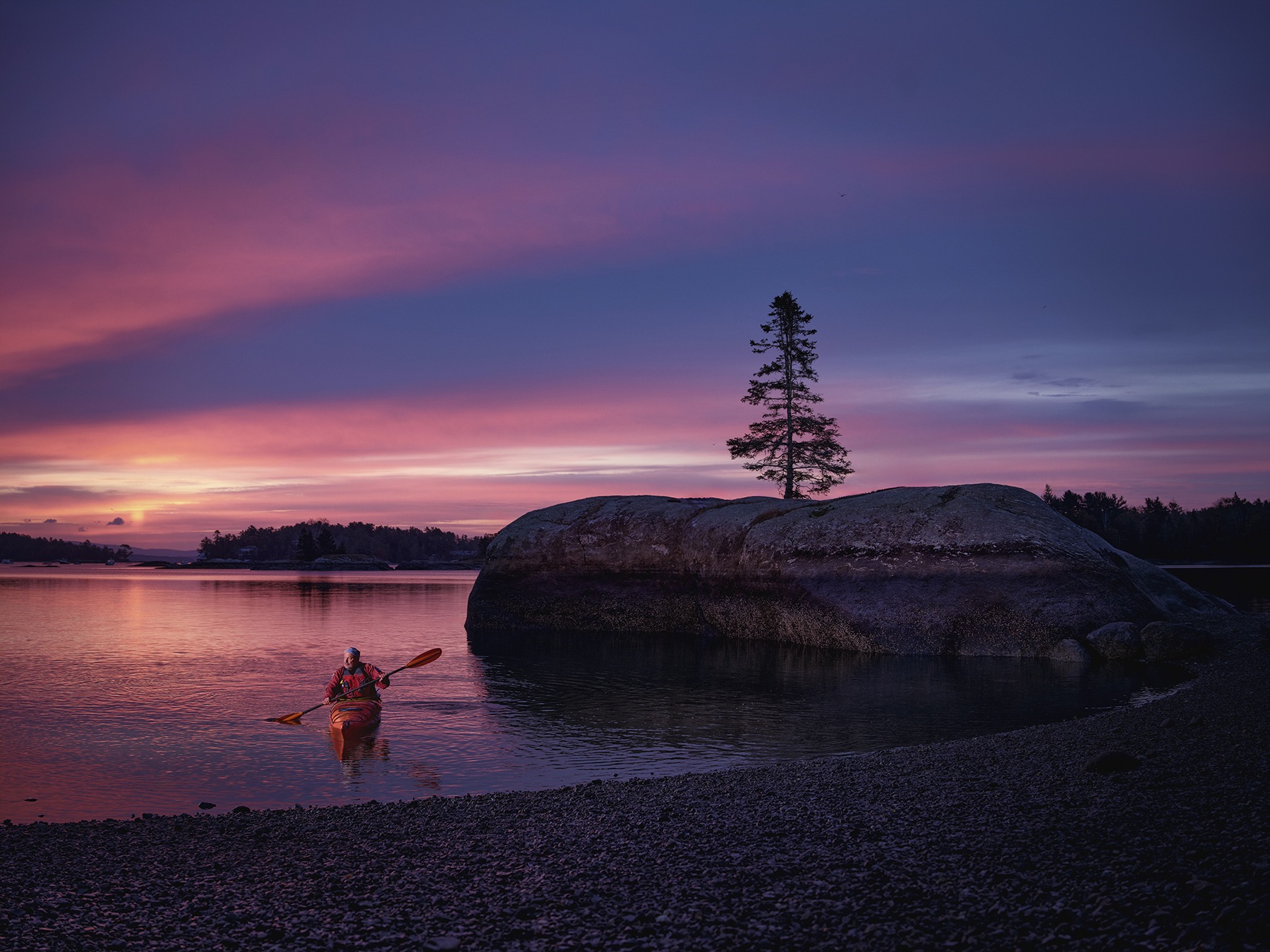 Person paddles a kayak on calm water at sunset near a rocky shoreline with a tree-covered rock formation under a colorful sky.