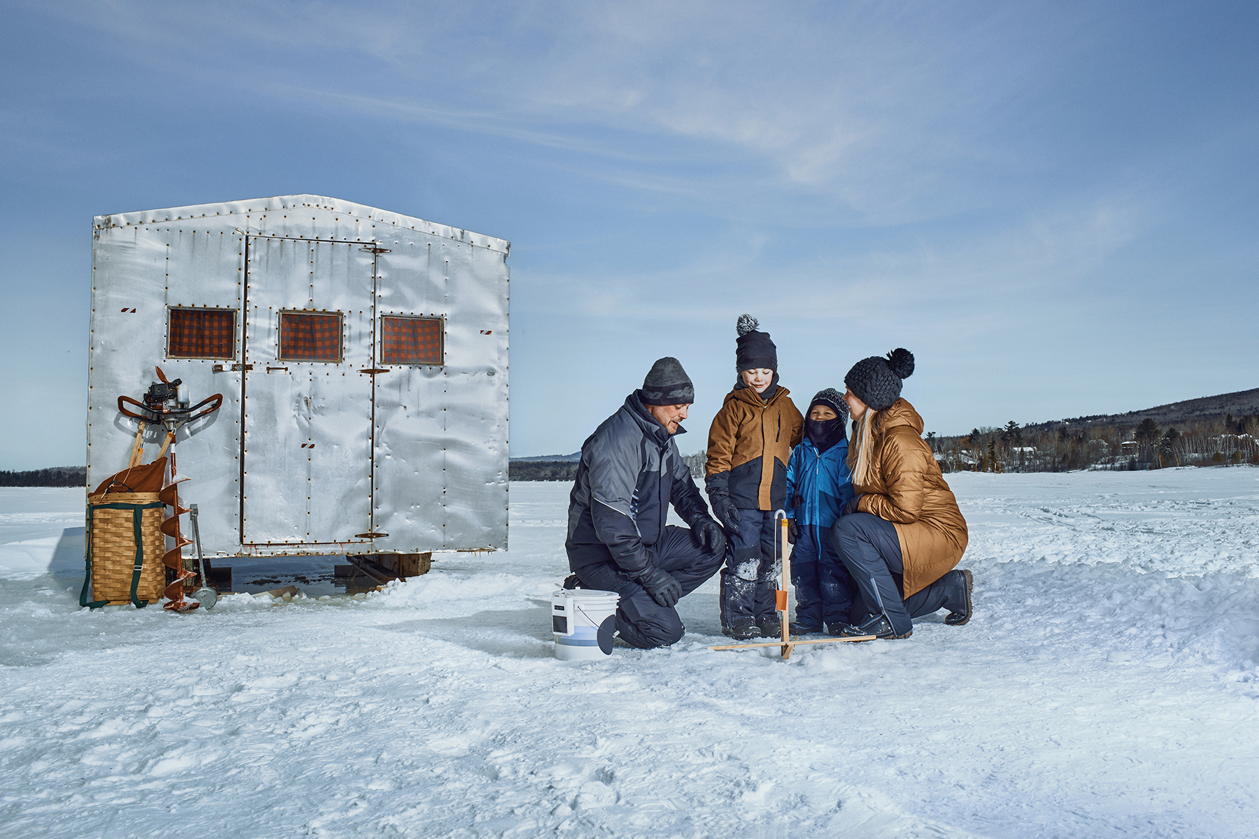 A family of four stands outside an ice fishing shack on a frozen lake on a bright day, enjoying the winter scenery and ice fishing.