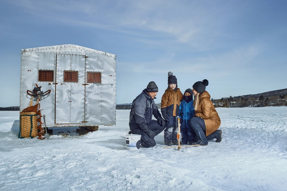 A family of four stands outside an ice fishing shack on a frozen lake on a bright day, enjoying the winter scenery and ice fishing.
