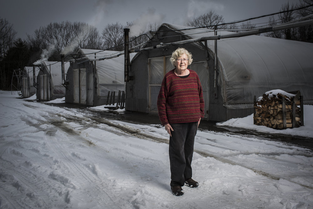 Full-length portrait of an older woman standing in front of greenhouses on a snowy day, with firewood stacked nearby.