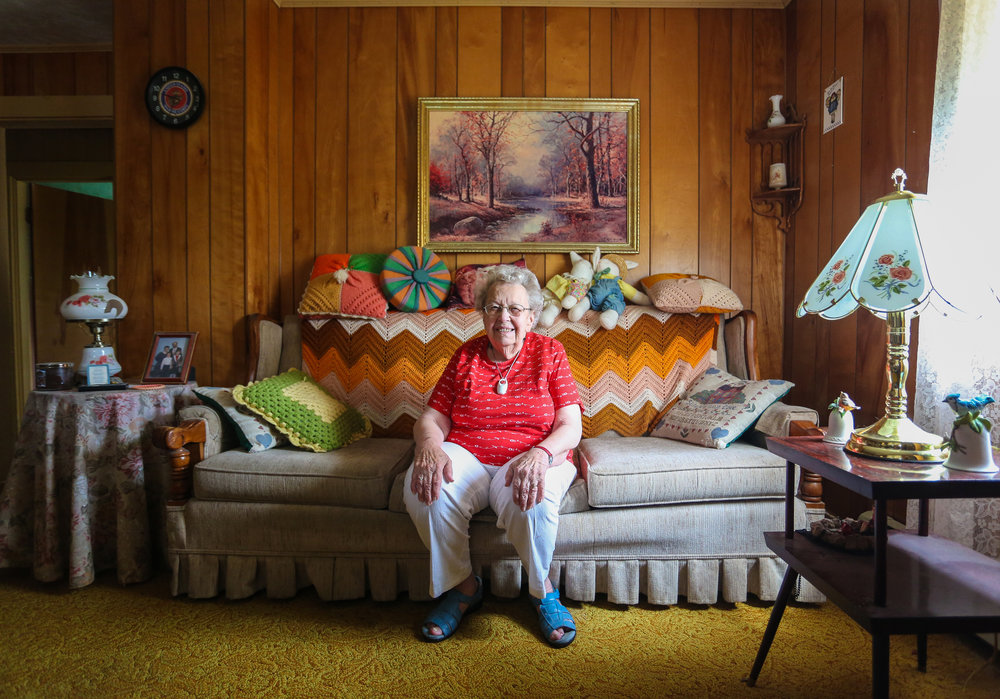 Elderly woman sits on a floral couch in a wood-paneled living room with crocheted pillows, a painting, and vintage lamps, smiling warmly.