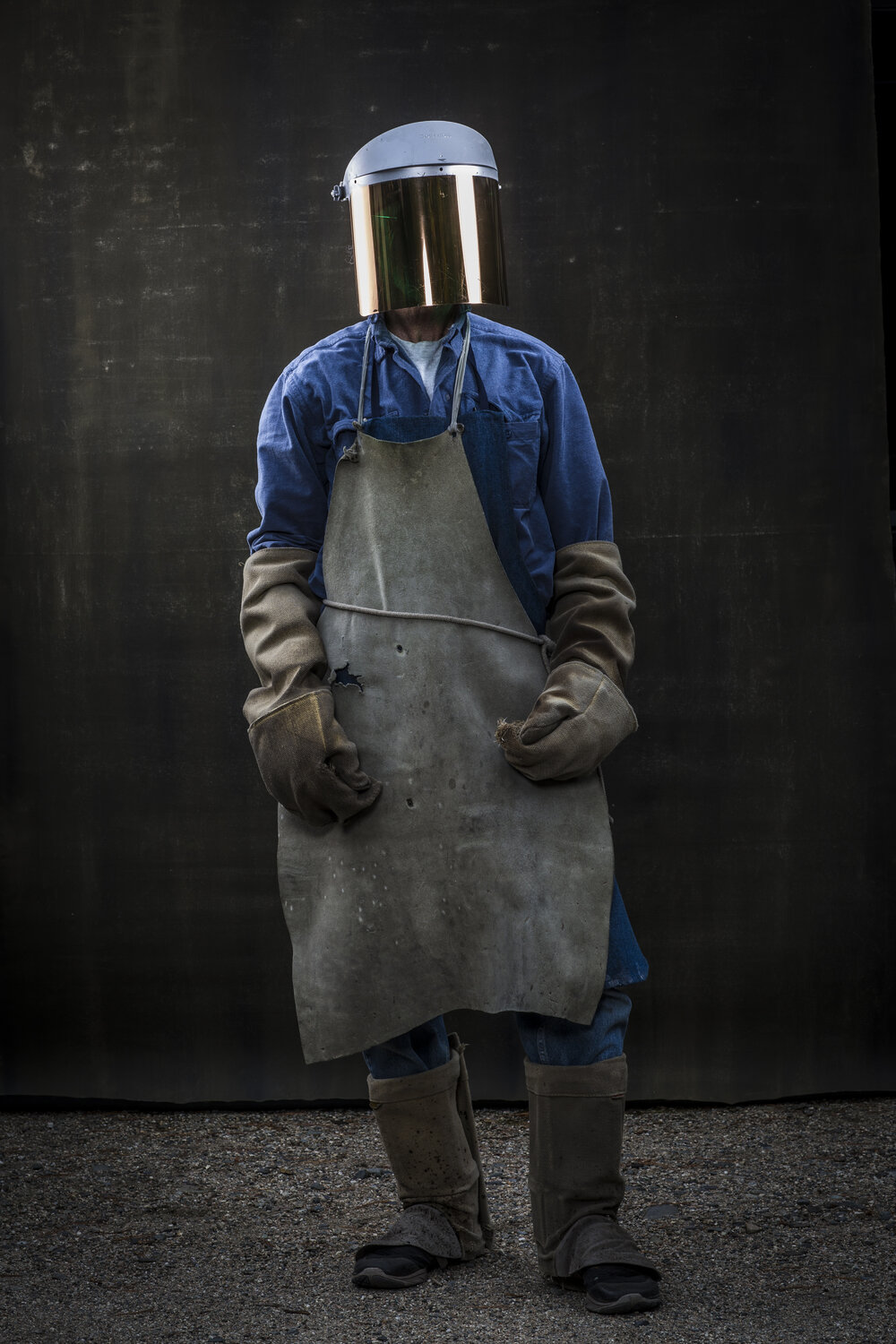 Person wearing a safety helmet, visor, apron, gloves, and boots stands in front of a dark background, facing the camera in a full shot.