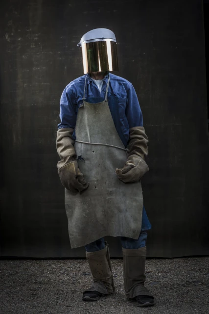 Person wearing a safety helmet, visor, apron, gloves, and boots stands in front of a dark background, facing the camera in a full shot.