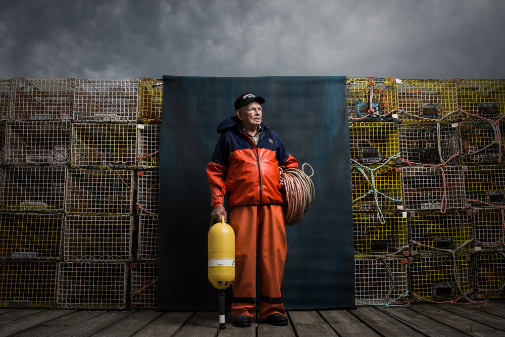 Elderly fisherman poses with orange waders, a yellow buoy, and coils of rope against a backdrop of stacked lobster traps under a cloudy sky.