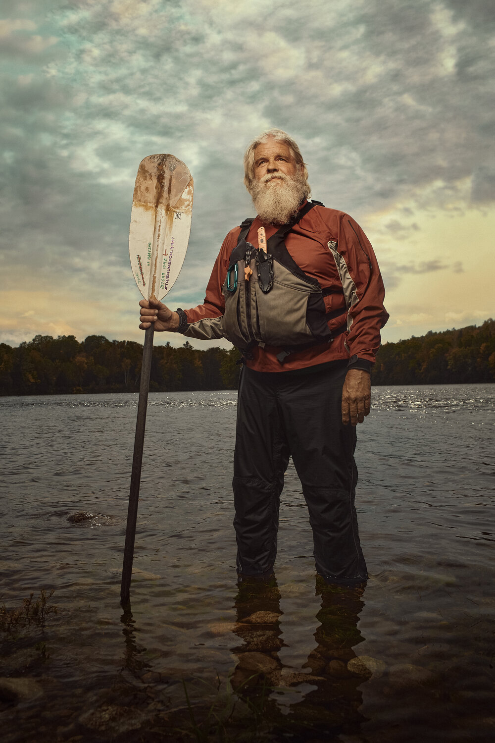 A bearded man in a red jacket stands knee-deep in a lake, holding an oar, with trees on the shoreline and a cloudy sky.