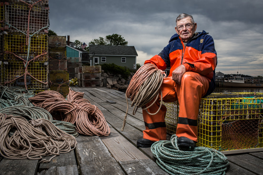 Elderly fisherman sits on a lobster trap on a wooden dock holding ropes, wearing an orange waterproof jacket and pants, with cloudy skies.