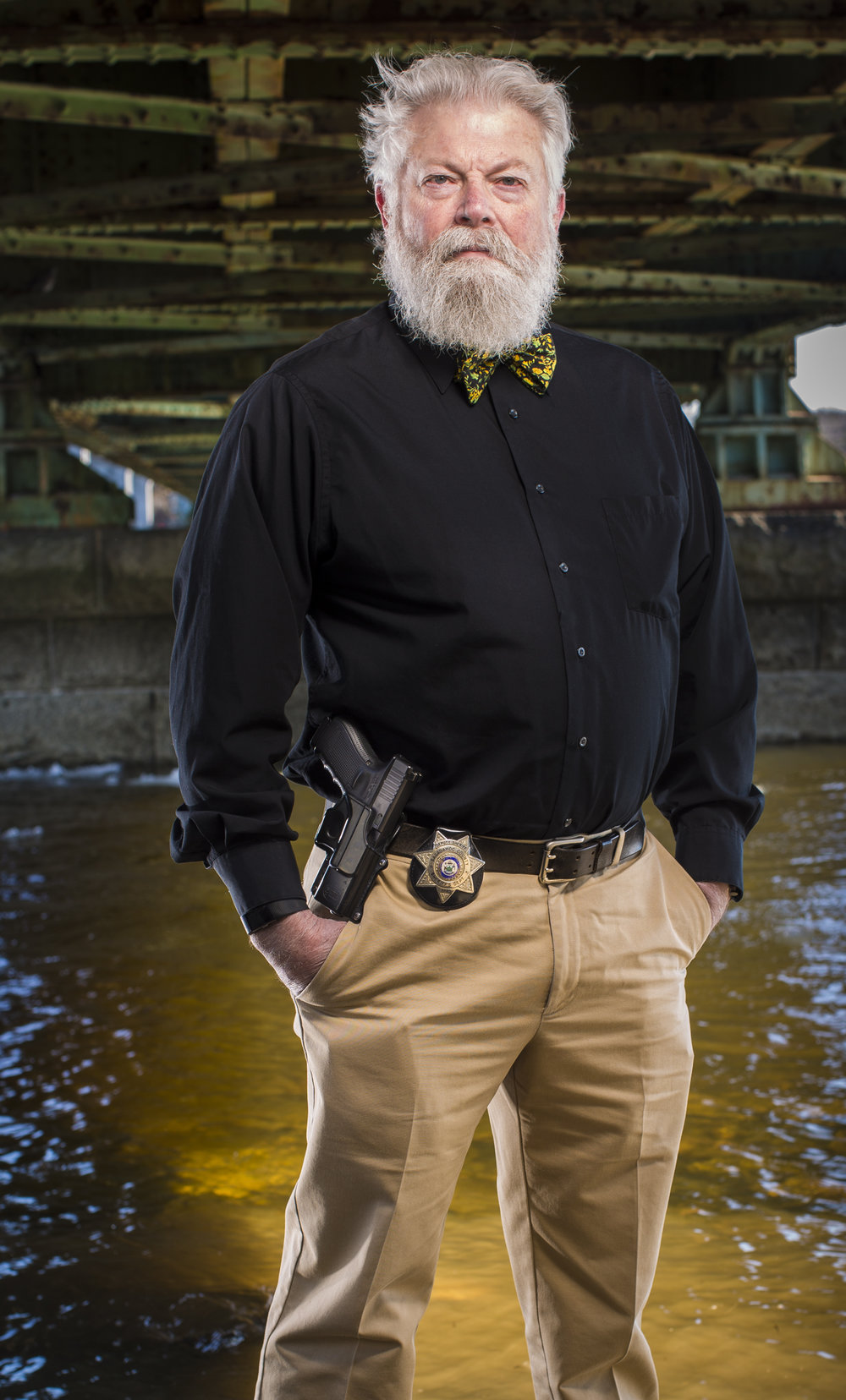 Man with white beard in black shirt, yellow bowtie, khaki pants, and badge stands under a bridge with a gun in a holster.