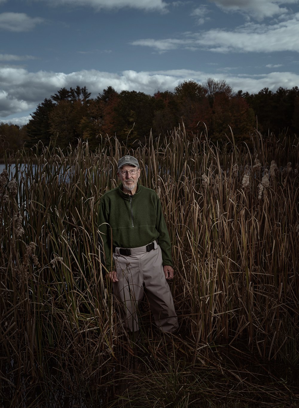 Portrait of an adult male standing in a field of tall grass by a lake, wearing outdoor casual clothing, under a cloudy sky.