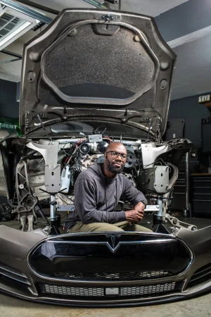Man sitting in front of the chassis of a deconstructed Tesla in a garage, wearing a gray hoodie and glasses, looking at the camera.
