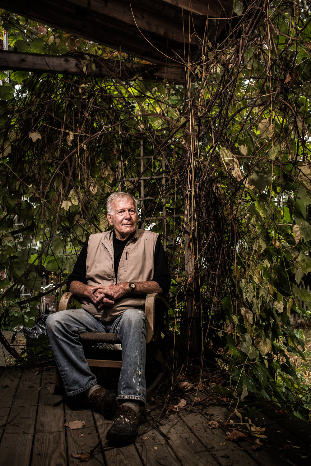 Older man with gray hair, wearing a vest and jeans, sits in a wooden chair on a rustic porch surrounded by vines and natural light.