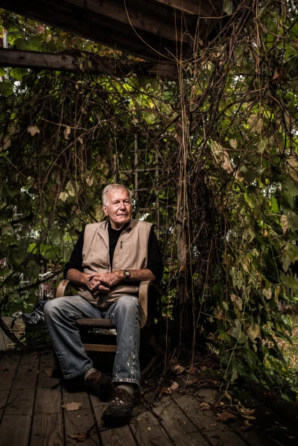 Older man with gray hair, wearing a vest and jeans, sits in a wooden chair on a rustic porch surrounded by vines and natural light.