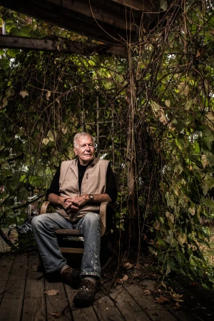 Older man with gray hair, wearing a vest and jeans, sits in a wooden chair on a rustic porch surrounded by vines and natural light.