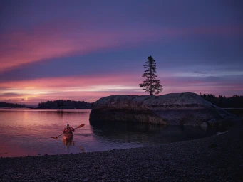 Person kayaking on calm water near a large rock with a single pine tree under a vibrant sunset sky