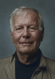 Portrait of an elderly man with short blonde hair wearing a dark turtleneck and tan jacket against a muted grey background.