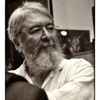 Black and white portrait of an adult male with a beard, wearing a light-colored shirt, with a blurred background featuring artwork.