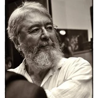 Black and white portrait of an adult male with a beard, wearing a light-colored shirt, with a blurred background featuring artwork.