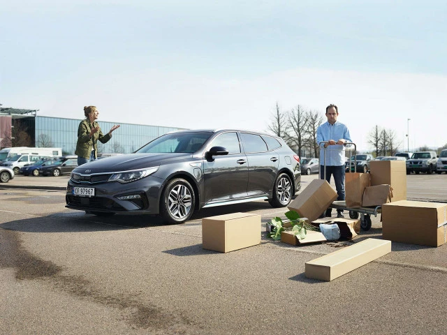 Grey Kia station wagon parked in a parking lot with a woman and a man standing nearby with moving boxes and a shopping cart.