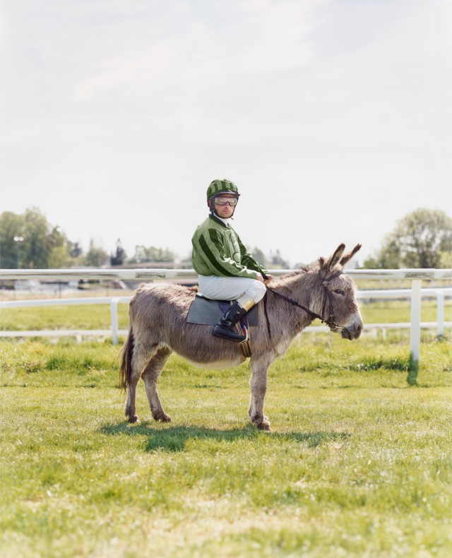 Jockey on a donkey at an outdoor race track; the man wears a green and black helmet and green striped jacket on a sunny day.