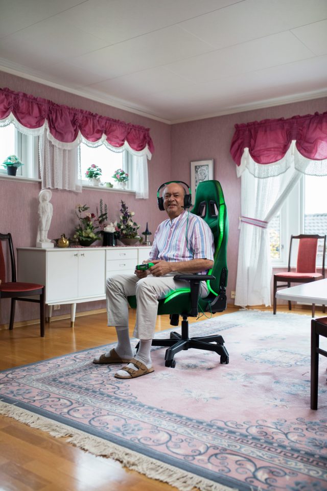 Elderly man wearing headphones, sitting in a green gaming chair with controller in his hands, inside a living room with a carpet.