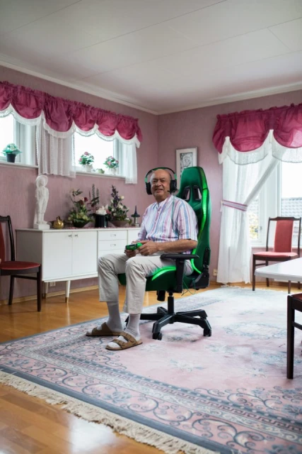 Elderly man wearing headphones, sitting in a green gaming chair with controller in his hands, inside a living room with a carpet.