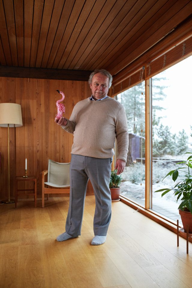 Mature man poses indoors holding a pink flamingo figurine, clad in beige sweater and gray pants, with a snow-covered landscape visible through window.