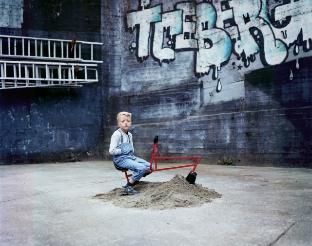 A young child in denim overalls sits on a toy excavator atop a sand pile outdoors, backed by a textured wall with graffiti art.