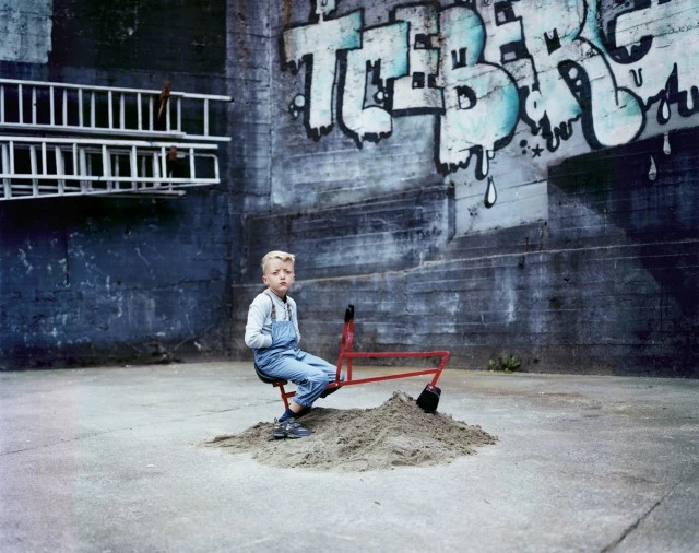 A young child in denim overalls sits on a toy excavator atop a sand pile outdoors, backed by a textured wall with graffiti art.