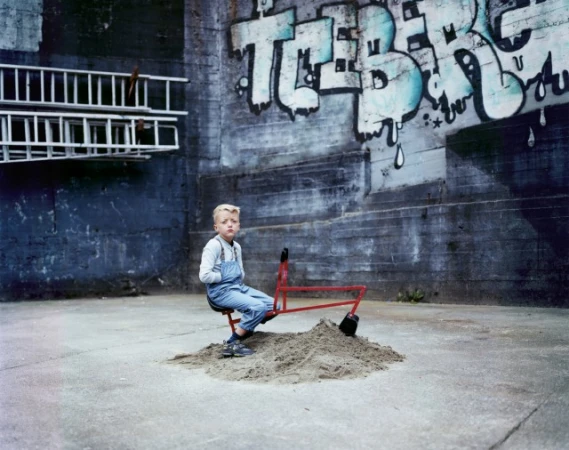 A young child in denim overalls sits on a toy excavator atop a sand pile outdoors, backed by a textured wall with graffiti art.