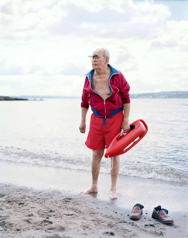 Elderly man in red jacket and shorts stands barefoot on beach with a lifeguard buoy. Ocean and cloudy sky backdrop on overcast day.
