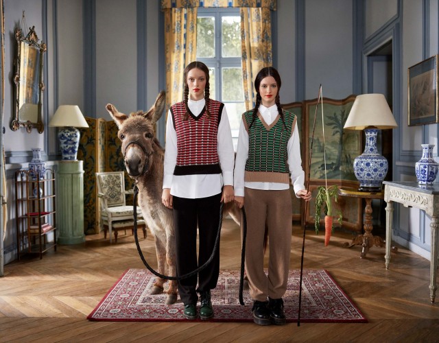 Two women in vests stand in an ornate room with a donkey on a leash, holding a carrot. Interior with hardwood floors and patterned rug.