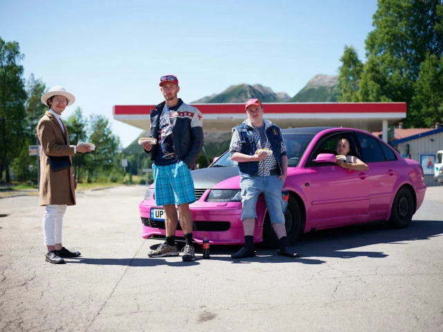 Three adult males and a woman pose with a pink car outside a gas station on a sunny day, sharing snacks near mountains.