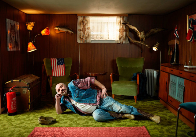 Man lounges on a green rug in a retro-styled room with wood paneling, vintage furniture, lamps, and decorative objects, under warm lighting.