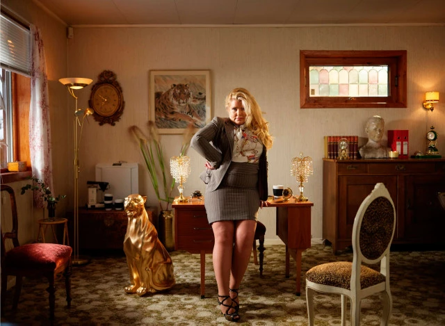 Woman in a grey skirt suit poses in a vintage-style room with gold accents, including a lion statue and ornate lamps, under warm lighting.