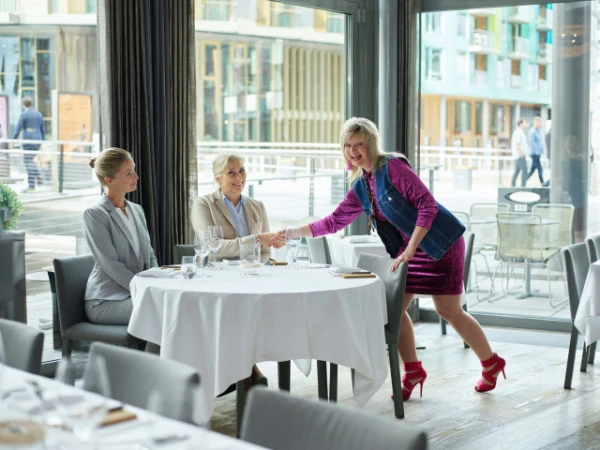 Three women in a restaurant shaking hands. Two seated at a table set for lunch, and one standing shaking the hand of a seated woman.