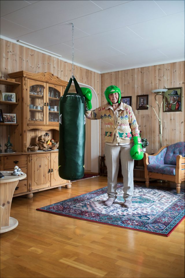 Elderly woman in boxing gear stands in a wood-paneled room with a punching bag, antique furniture, and a patterned rug.