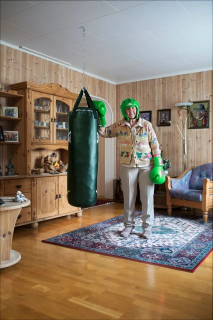 Elderly woman in boxing gear stands in a wood-paneled room with a punching bag, antique furniture, and a patterned rug.