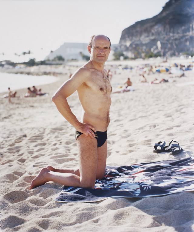 A bald mature man kneeling on a patterned beach towel on sand with mountains in the background by Morten Borgestad