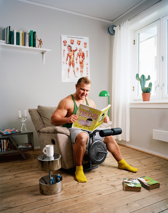 Muscular man in tank top sits in armchair reading weight-training book indoors with exercise equipment and books on the floor.