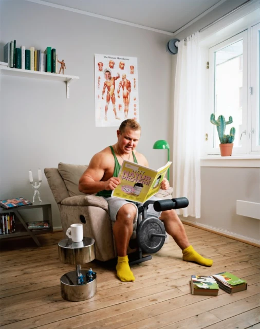 Muscular man in tank top sits in armchair reading weight-training book indoors with exercise equipment and books on the floor.