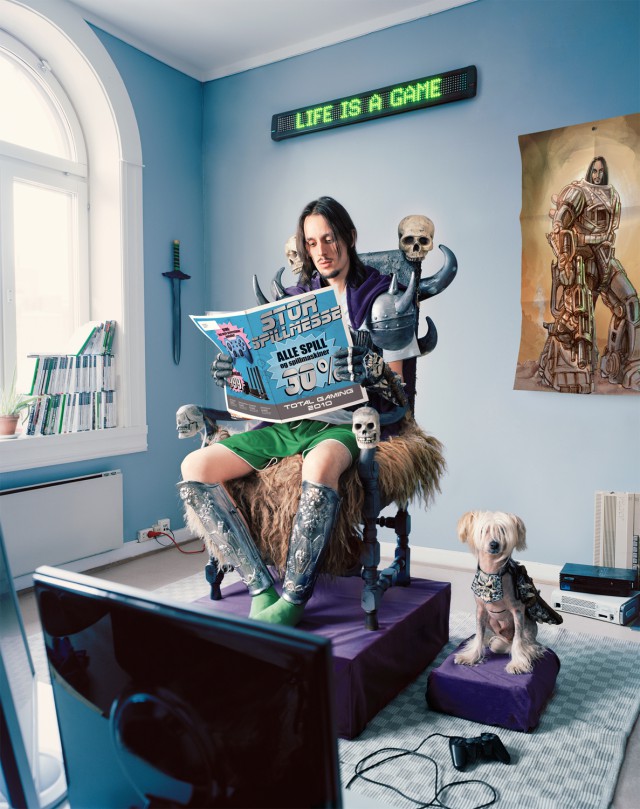 Man in fantasy costume reading in a themed room with a dog. Gamer aesthetic with comic book, skull decor, and a digital wall display.