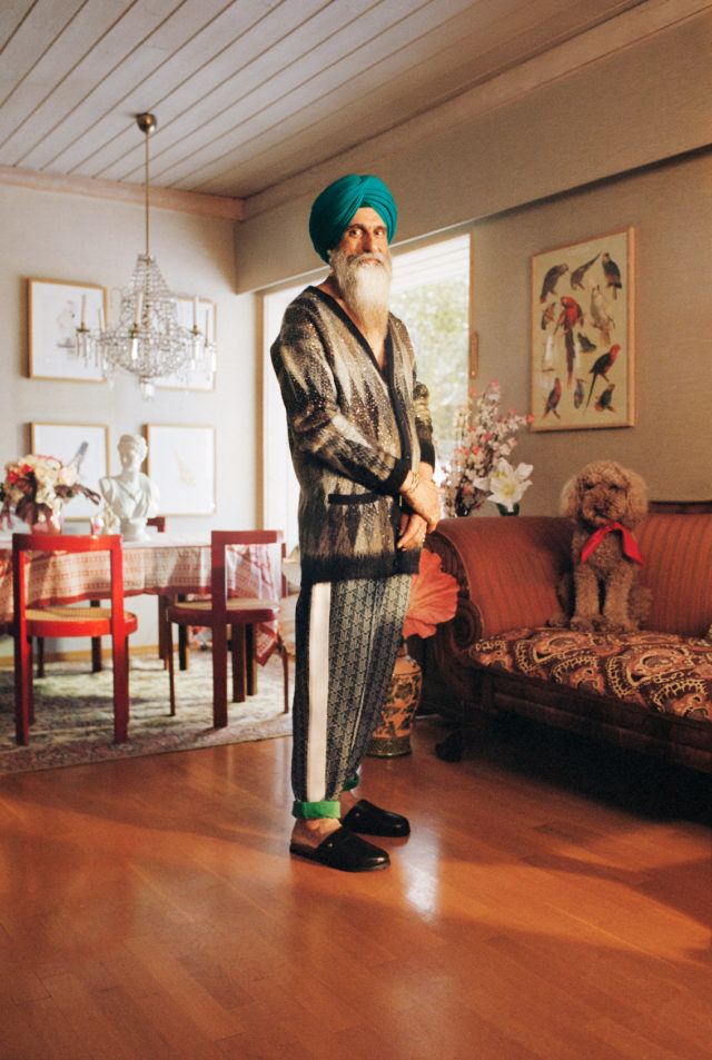 Full shot of a bearded man in a turban standing in an ornate living room next to a poodle on a couch. Hardwood floors.